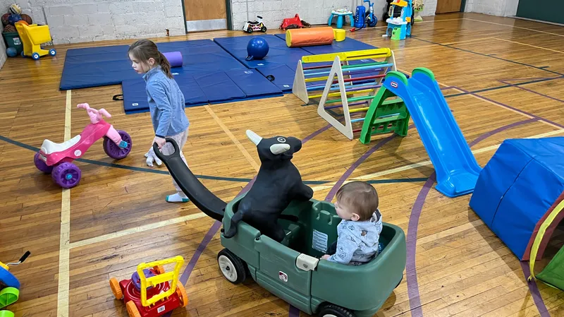 Picture of a child in a wagon being pulled by another child at Tot Spot Lancaster. There is a toy bull in the wagon and gym matts and climbing toys are in the background.